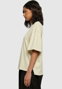 Beige bavlněné tričko s krátkým rukávem, volný střih, kulatý výstřih a boční rozparky. Model stojí v profilu.