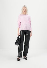 Model staat tegen een egale achtergrond, draag een lichtroze gebreide trui, zwarte leren broek en houdt een zwarte handtas vast.