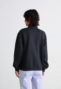 Sweatshirt noir à col montant et épaules tombantes, avec une texture douce et une coupe décontractée, associé à un pantalon mauve clair.