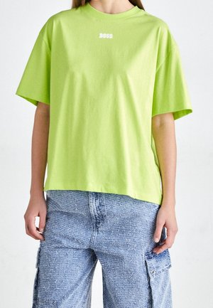 Osoba ubrana w jaskrawozielony oversize T-shirt z tekstem "BOSS" oraz niebieskie teksturowane spodnie cargo, stojąca na jednolitym tle.