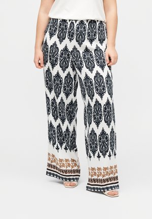 Kobieta w białych spodniach o szerokich nogawkach z czarnymi i brązowymi misternymi wzorami oraz białych sandałach, stojąca na jednolitym tle.