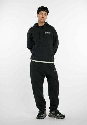 Jonge persoon staat in een zwart New Balance hoodie en trainingsbroek met zwart en grijs sneakers tegen een effen witte achtergrond.