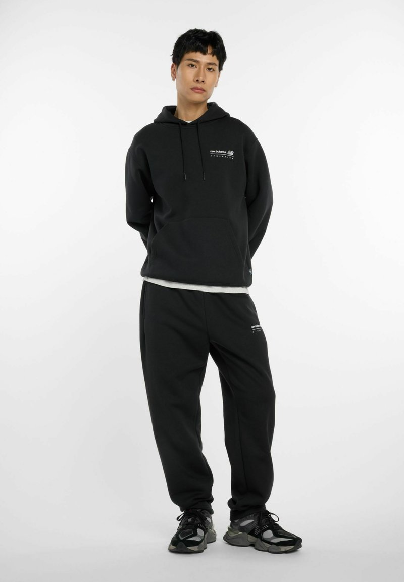 Jeune personne debout portant un hoodie noir New Balance et un pantalon de jogging, avec des baskets noires et grises, devant un fond blanc uni.