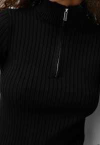 Černý vroubkovaný pletený top s vysokým límcem a stříbrným zipem. Tkanina má vertikální pruhy, což poskytuje texturovaný vzhled.