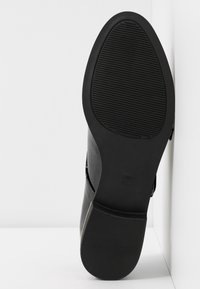 Chaussure en cuir noir avec une surface lisse et une semelle en caoutchouc dotée de rainures horizontales pour l'adhérence. La chaussure présente un design de bout arrondi.