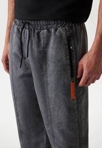 Joggers ajustados en gris, hechos de una tela suave, con una cinturilla elástica, bolsillo lateral con cremallera y un parche de logo naranja. Superficie texturizada.