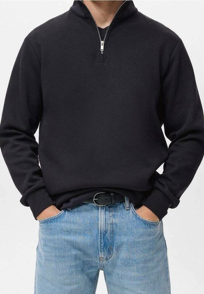 Homme portant un sweat-shirt noir à fermeture éclair jusqu'au quart, rentré dans un jean bleu clair avec une ceinture noire, les mains dans les poches.