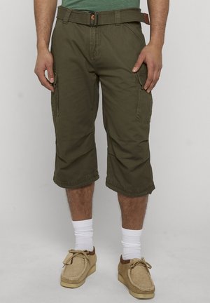 Mand iført olivengrønne cargobukser, brun bælte, hvide sokker og beige afslappede sko, stående mod en ensfarvet baggrund.