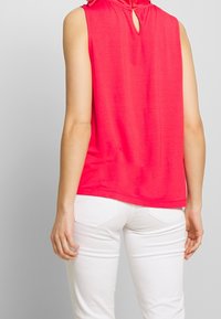 Haut sans manches en tissu rose vif avec une découpe en forme de trou à l'arrière, porté avec un jean blanc ajusté. Texture lisse, design décontracté.