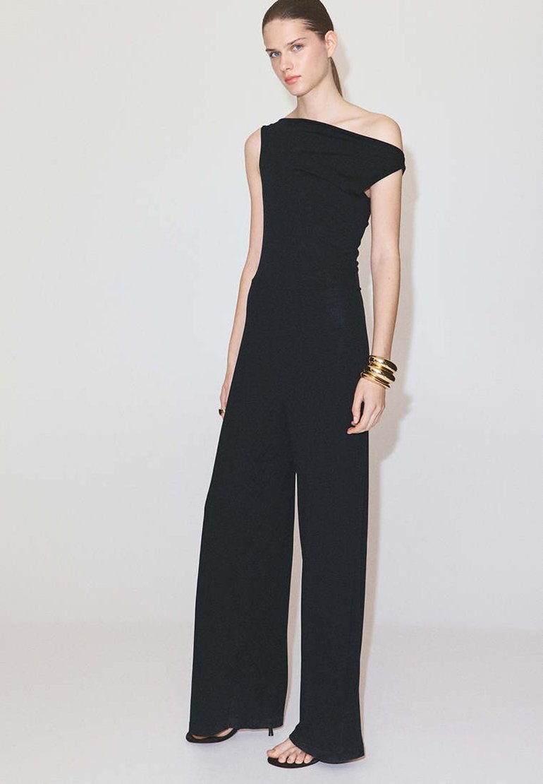 Femme portant une combinaison noire asymétrique à larges jambes et bracelets dorés, debout devant un fond clair uni.