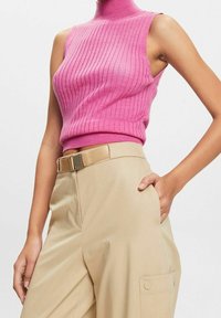 Vaate ilman hihoja, vaaleanpunainen ribbikuvioinen turtleneck yllään, beige korkeavyötäröiset housut, joissa on vyö ja sivutasku, käsi taskussa asennossa.