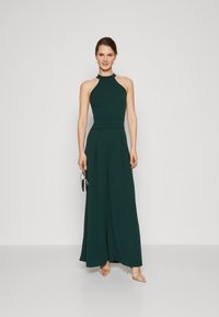 Halter-neck jumpsuit i dyp grønn stoff, med en tettsittende bodice, plissert bredbeint bukser, og en jevn tekstur.
