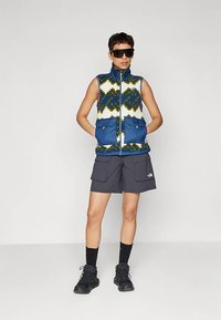 Person som har på seg et mønstret blågrønt og hvitt ermeløst vest med store frontlommer, mørke cargo-shorts, svarte sokker, svarte tursko og store solbriller.