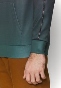 Felpa con cappuccio in tessuto verde scuro con tasca a marsupio. Presenta polsini a contrasto e una cucitura con testo in rosso e bianco.