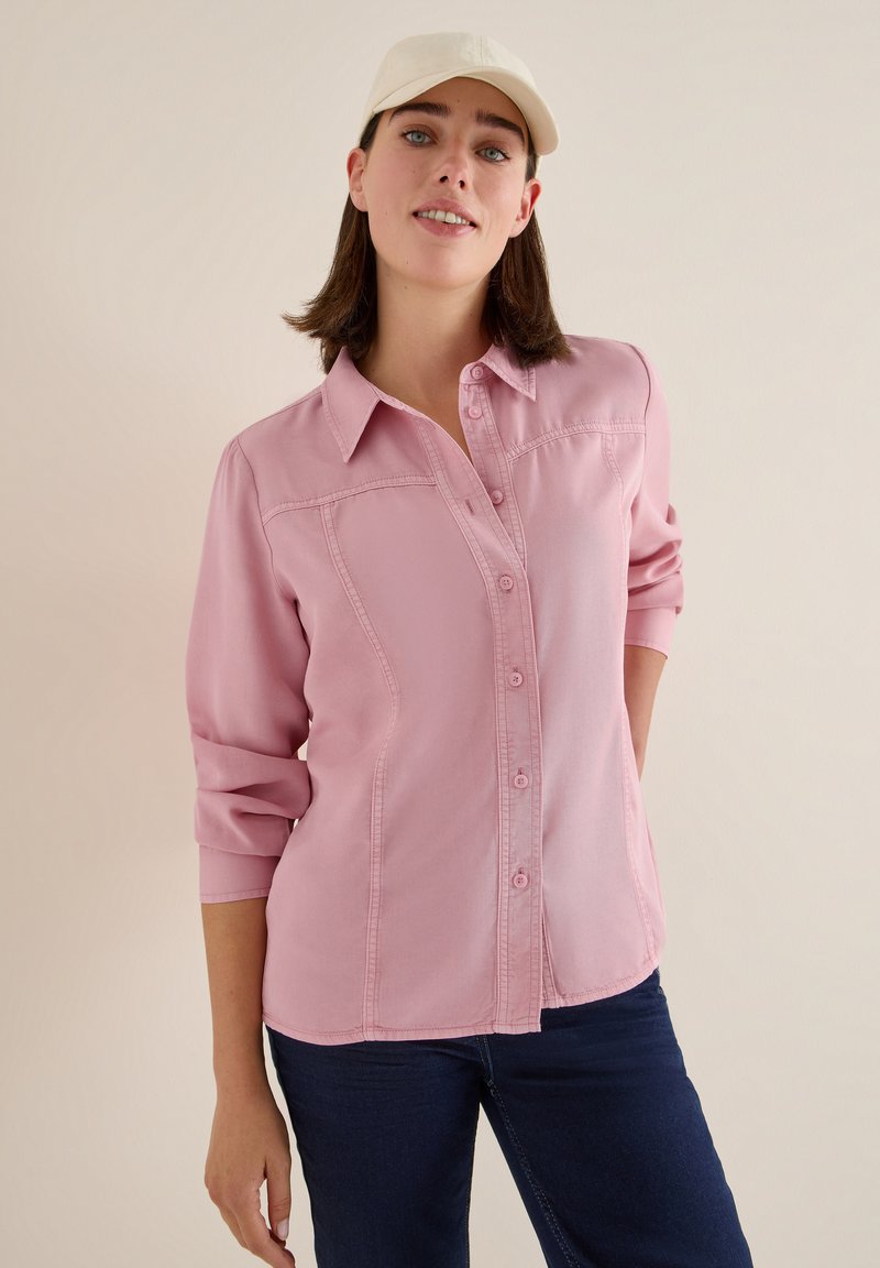 Helles rosa Langarmhemd mit Knopfleiste, klassischem Kragen und hochgekrempelten Ärmeln, mit Seiten Nähten und subtiler Textur. Kombiniert mit dunklen Jeans.