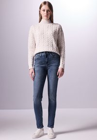 Pull en maille torsadée crème avec col montant, jeans skinny bleus et baskets blanches. Textures douces et coupe ajustée soulignent un look décontracté.