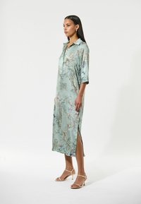 Fließendes mintgrünes Kleid mit Blumenmuster, Kragen, halblangen Ärmeln, seitlichen Schlitzen, aus glattem Stoff.