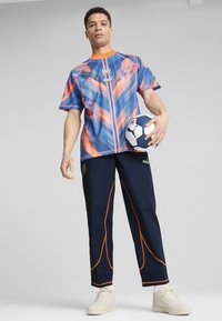 Sporta apģērbs, kas sastāv no zilas un oranžas rakstotas īsām piedurknēm krekla un tumši zilām biksēm ar oranžiem akcentiem, tur rokā futbola bumbu.