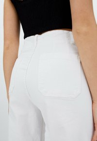 Persona que lleva pantalones blancos de tiro alto con bolsillos traseros y un top negro recortado, visto desde la cintura hacia abajo por detrás.
