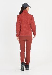 Röd idrottsjacka med hög krage och långa ärmar, kombinerad med bruna avsmalnande byxor och svarta vandringskängor. Slät textur, minimalistisk design.