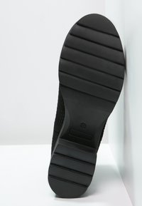 Czarna podeszwa buta z równoległymi rowkami, z chropowatym boku. Materiał przypomina gumę, zaprojektowany z myślą o przyczepności i trwałości.