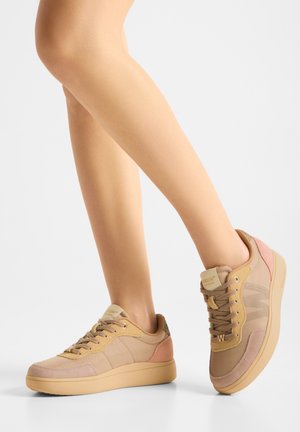 Piernas con zapatillas casuales beige y rosas con cordones beige, de pie sobre una superficie blanca con un fondo blanco liso.
