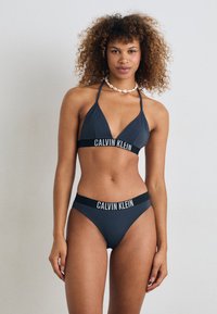Set bikini grigio scuro con top a halter e pantaloni alti, caratterizzato da una cintura nera a contrasto con la scritta "CALVIN KLEIN" stampata in bianco.