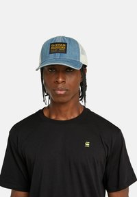 Gorra de mezclilla y malla con el frente azul y la parte trasera blanca, que presenta una etiqueta rectangular negra con texto amarillo. Camiseta negra debajo.
