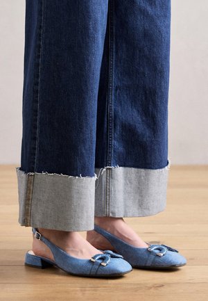 Dolne części nóg w szerokich, podwiniętych ciemnoniebieskich jeansach oraz jasnoniebieskich butach typu slingback z ozdobnymi klamrami na drewnianej podłodze.