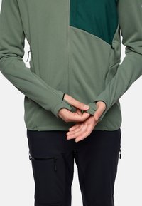 Giacca verde con zip, dotata di una tasca sul petto verde scuro e aperture per i pollici sulle maniche; indossata sopra pantaloni neri con tasche laterali con zip.