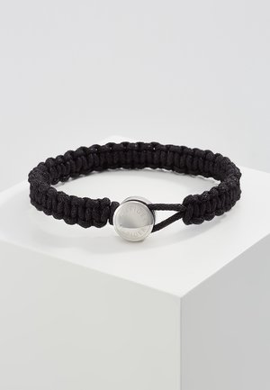 Schwarzes geflochtenes Cord-Armband mit rundem silbernem Verschluss, der mit "Hilfiger" graviert ist, präsentiert auf einem weißen Sockel vor einem schlichten Hintergrund.