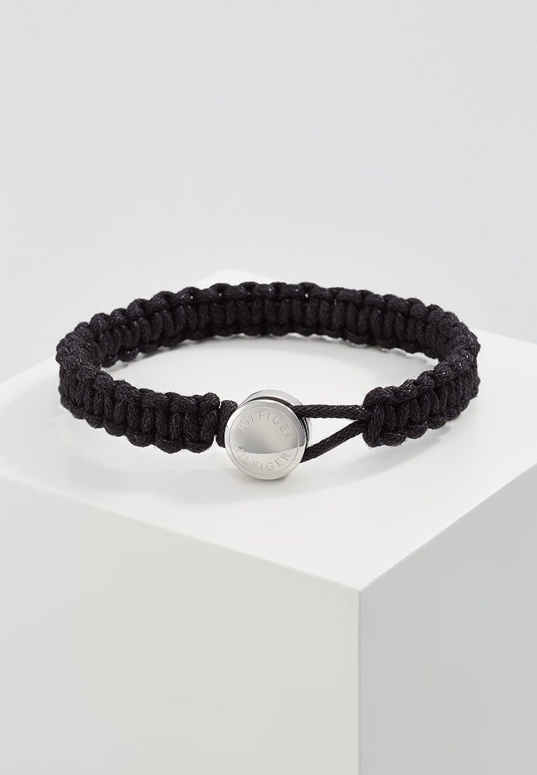 Bracelet en cordon noir tressé avec un fermoir rond en argent gravé de "Hilfiger", présenté sur un socle blanc contre un fond uni.