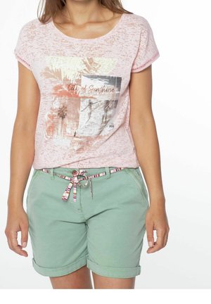 Kvinde iført pastelrosa grafisk t-shirt og grønt opsmøgede shorts med en stribet stofbælte mod en hvid baggrund.
