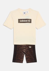 Zestaw dziecięcy Adidas składający się z beżowej koszulki z kamuflażowym logo oraz brązowych spodni krótkich z wzorem i czarnymi bocznymi paskami.