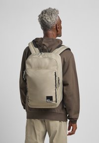 Mochila de nylon beige con un bolsillo frontal con cremallera, acentos negros y logotipo. Correas ajustables acolchadas y forma rectangular plana.