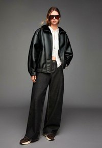 Črna usnjena jakna iz umetnega usnja z visokim ovratnikom, v kombinaciji s širokimi temno denim hlačami in rjavimi športnimi copati. Ima rebrasto strukturo na rokavih.