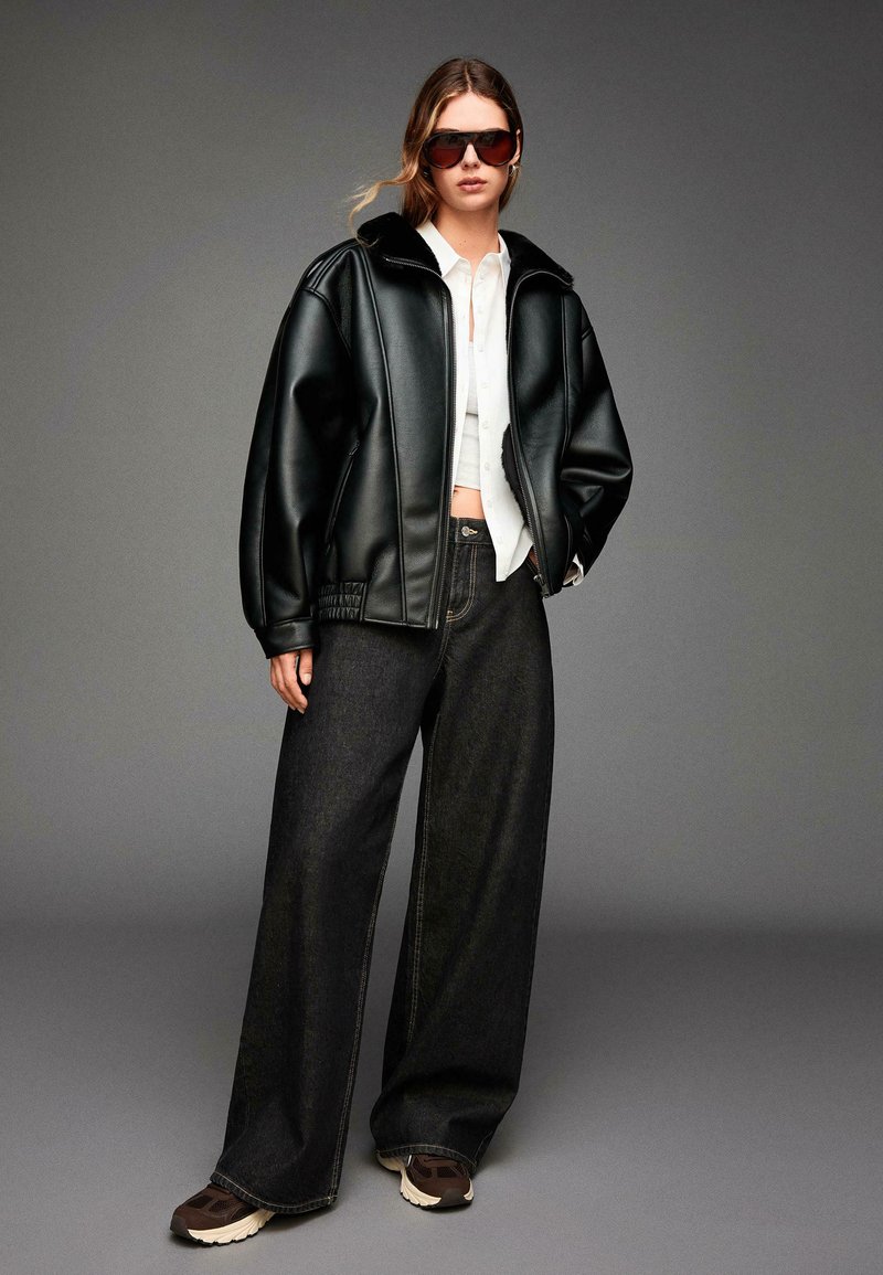 Črna usnjena jakna iz umetnega usnja z visokim ovratnikom, v kombinaciji s širokimi temno denim hlačami in rjavimi športnimi copati. Ima rebrasto strukturo na rokavih.