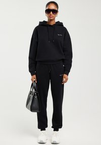 Naine, kes kannab musta kapuutsiga pusa ja spordipükse, valged tossud, päikeseprillid ning hoiab käes musta käekotti tavalise tausta ees.