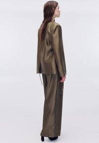 Olivengrønt jakkesæt lavet af glat, skinnende stof. Skræddersyet jakke med bagventil, parret med løstsiddende, vidde bukser og høje hæle.