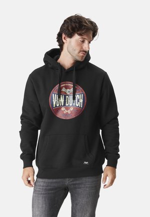 Homme portant un sweat à capuche noir avec un logo circulaire rouge et blanc "Von Dutch", associé à un jean gris, regardant légèrement vers le bas et sur le côté.