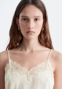 Cremfarbene Camisole mit Spitzenrand und dünnen Trägern, die an der Ausschnittkante mit filigranen Blumenmustern verziert ist. Weiche, glatte Textur.