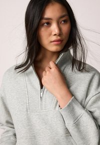 Grå sweater med høj krave og lynlåslukning. Har ribbede mansjetter og et tekstureret stof, med en afslappet pasform.