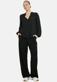 Woman wearing a black button-up blouse with lace detail and loose black trousers, standing with hands in pockets and white sneakers.