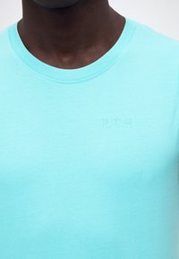 Κοντινό πλάνο ενός ατόμου που φοράει ένα ανοιχτό μπλε t-shirt με στρογγυλή λαιμόκοψη και αχνό κείμενο "1962 Tom Tailor" στο αριστερό στήθος.