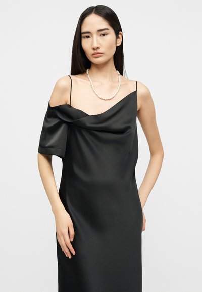 Γυναίκα με μακριά σκουρόχρωμα μαλλιά, που φοράει ένα μαύρο σατέν φόρεμα με ακάλυπτους ώμους και ένα μονόκλωνο λευκό περιδέραιο από πέρλες, στέκεται μπροστά σε απλό φόντο.