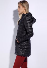 Zwarte pufferjack met een capuchon, voorzien van een quiltpatroon, ritszakken en een gladde, glanzende textuur. Draagt over rode broek.