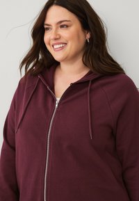 Burgunderfarbener Zip-Hoodie mit Frontreißverschluss, Kapuze mit Kordelzug und weicher Textur. Verfügt über gerippte Bündchen und eine lockere Passform.