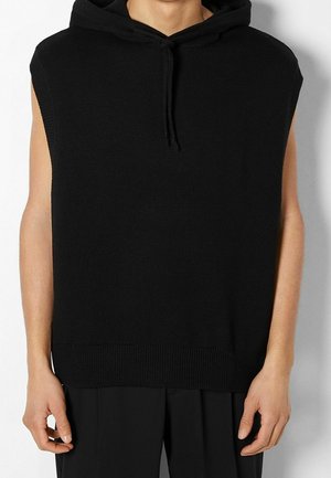 Haut en maille noir sans manches à capuche porté par une personne les mains détendues le long du corps, assorti d'un pantalon noir sur un fond uni.