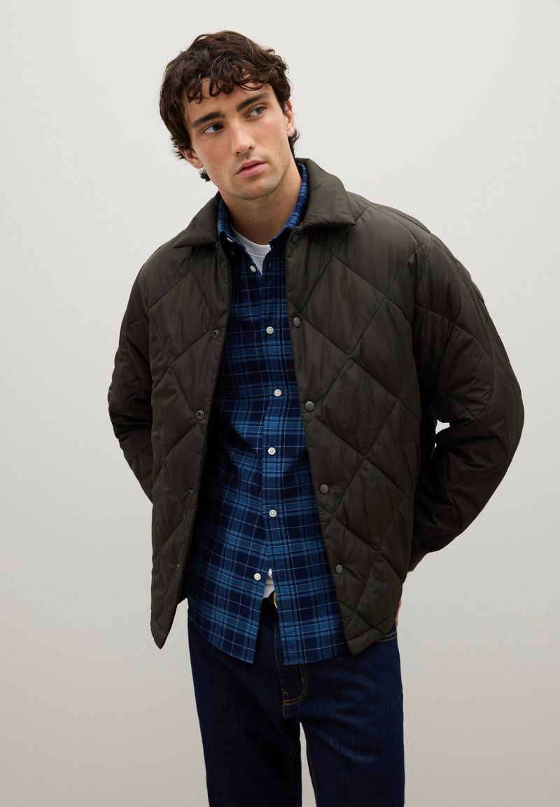 Giacca trapuntata verde scuro con colletto, dotata di bottoni a pressione e motivi a rombi, indossata sopra una camicia a quadri blu.