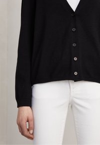 Cardigan noir avec un col en V, doté d'une fermeture à boutons et de poignets côtelés. Assorti à un jean blanc ajusté. Texture douce, coupe décontractée.